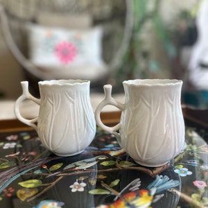 May include: Two white ceramic mugs with a leaf design. The mugs have a handle on the side and a spout on the top. The mugs are sitting on a black tray with a floral design.