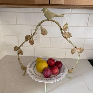 May include: A decorative metal fruit stand with a bird and pear design. The stand holds a plate with a yellow pear and red plums. The stand has a light green and gold finish, with a white and blue floral plate.
