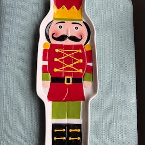 May include: A white ceramic serving platter in the shape of a toy soldier nutcracker. The platter features a red jacket, green pants, and black boots. The nutcracker has a black mustache, a gold crown, and is decorated with yellow accents.