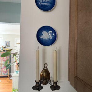 May include: Two circular blue and white porcelain plates featuring relief images of a swan and a bird, mounted vertically above a brass double-arm wall sconce holding two off-white taper candles with a metallic finish on the candle holders.