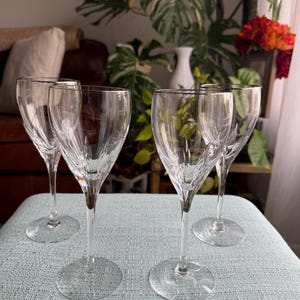 May include: Four clear crystal wine glasses with etched designs and silver rims are arranged on a light blue surface. The glasses have long stems and round bases. The background includes a couch, plants, and a window.