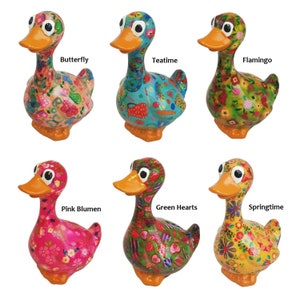 Op de afbeelding: Zes kleurrijke keramische eendjes met verschillende bloemenpatronen. De eendjes kijken allemaal naar voren met hun snavels open. De patronen omvatten vlinders, theekopjes, flamingo's, roze bloemen, groene harten en een lenteontwerp.