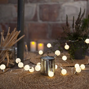 Op de afbeelding: Een slinger van warmwitte gloeilampen op een tafel. De lichten zijn gewikkeld rond een conservenblik, een potplant en een geweven placemat. Broodstengels in een glazen pot en kaarsen creëren een gezellige sfeer.