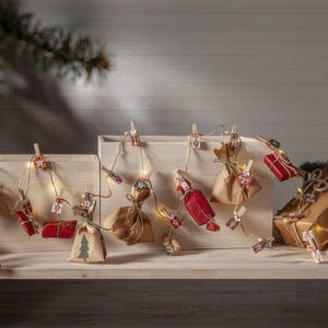 Könnte beinhalten: Adventskalender mit kleinen verpackten Geschenken, roten und braunen Papierpaketen und einer Lichterkette. Einige Geschenke sind rot, andere braun, und einige haben ein kleines Weihnachtsbaum-Design. Auf einem Holzregal mit Tannenzweigen.