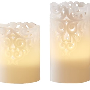 Op de afbeelding: Twee witte kaarsen in pilaarvorm met een decoratief, kant-achtig ontwerp. De kaarsen zijn aangestoken en geven een warm licht.