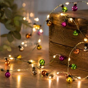 Könnte beinhalten: Eine dekorative Lichterkette mit kleinen, runden, bunten Glühbirnen in den Farben Grün, Lila, Orange und Silber. Die Lichter sind durch einen dünnen, flexiblen Draht verbunden und strahlen ein warmes Licht auf die Oberfläche ab.