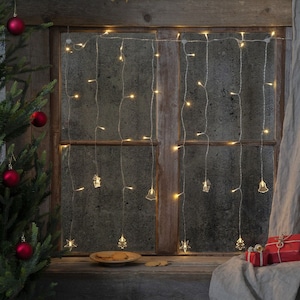 Op de afbeelding: Een rustiek houten raam met een string van warm witte kerstverlichting met kleine decoratieve ornamenten die eraan hangen. Het raam is versierd met een rood-wit geruit gordijn en een kerstboom met rode kerstballen.