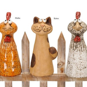 Könnte beinhalten: Drei Keramikfiguren eines Hahns, einer Katze und einer Henne, die auf einem Holzzaun stehen. Der Hahn ist orange und braun mit einem roten Kamm, die Katze ist braun und weiß mit schwarzen Flecken und die Henne ist weiß mit braunen Flecken. Die Figuren schauen alle nach oben.