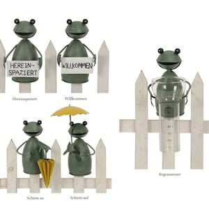 Könnte beinhalten: Vier grüne Froschfiguren mit verschiedenen Accessoires, die an einem weißen Lattenzaun befestigt sind. Die Accessoires umfassen ein Schild mit der Aufschrift "Herein-Spaziert", ein Schild mit der Aufschrift "Willkommen", einen Regenschirm und ein Regenmessgerät.