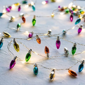 Könnte beinhalten: Eine Lichterkette mit dekorativen LED-Leuchten, die kleine, mehrfarbige Lampenabdeckungen in den Farben Pink, Blau, Grün, Orange und Silber aufweist. Die Lichter sind mit einem dünnen, flexiblen silbernen Draht verbunden und leuchten warm.