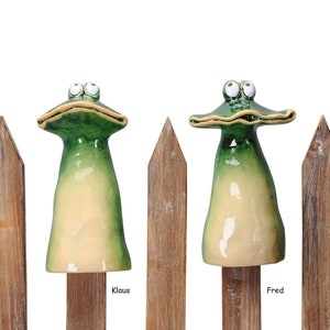 Könnte beinhalten: Zwei grüne Keramik-Froschfiguren mit großen Augen und offenem Maul sitzen auf Holzlatten. Der Frosch links ist mit "Klaus" und der Frosch rechts mit "Fred" beschriftet.