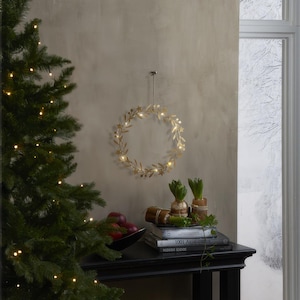 Könnte beinhalten: Ein goldfarbener, blattförmiger Kranz mit kleinen Lichtern hängt an einer Wand. Ein Weihnachtsbaum mit Lichtern befindet sich links. Ein schwarzer Tisch enthält Bücher, Äpfel und Pflanzen. Ein Fenster mit verschneiter Aussicht befindet sich rechts.