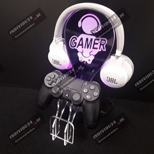 Könnte beinhalten: Ein weiß-schwarzes Gaming-Headset mit Mikrofon steht auf einem durchsichtigen Acryl-Ständer mit zwei schwarzen Videospiel-Controllern. Der Ständer hat eine durchsichtige Acrylplatte mit dem Wort "GAMER" und einem stilisierten Gamer-Symbol.