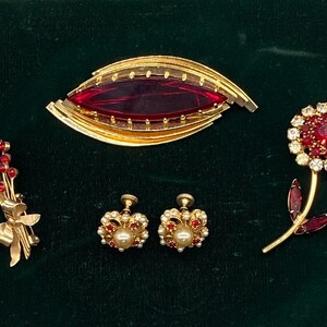 Puede incluir: Una colección de joyería en tono dorado que incluye un broche grande en forma de marquesa con una piedra roja central, un broche floral con cristales rojos y transparentes, un broche de ramo con detalles rojos y un par de pendientes de tornillo con perlas.