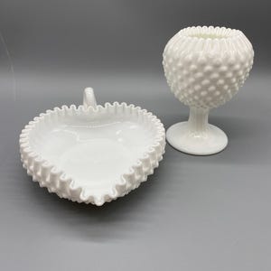 Puede incluir: Dos piezas de vidrio de leche Fenton hobnail blancas y vintage. Una es una fuente en forma de corazón con borde ondulado y asa. La otra es un jarrón hobnail con base de pedestal y borde superior ondulado. Ambas piezas están sobre un fondo gris.