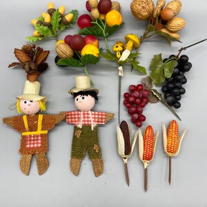 May include: A collection of autumn-themed decorative items including two fabric scarecrow figures with straw hats, artificial fruit and nut clusters, a bunch of dark grapes, a cluster of yellow mushrooms, and three individual ears of decorative corn.