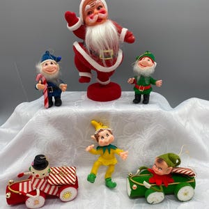 May include: A collection of five vintage-style Christmas ornaments featuring a Santa Claus figure, two standing elves, one elf in a yellow outfit, a snowman driving a red car with a gift, and an elf driving a green car with a candy cane.