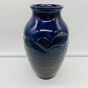 May include: A handcrafted ceramic vase with a dark blue glaze. The vase features a textured design on the upper portion and a gradient from dark blue to brown. The vase has a rounded body and a narrow neck.