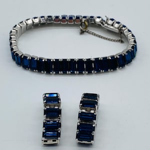 Könnte beinhalten: Ein Vintage-Schmuckset mit einem Armband und passenden Ohrringen. Das Armband und die Ohrringe haben eine silberfarbene Metalleinfassung und sind mit rechteckigen, tiefblauen Edelsteinen verziert. Das Set wird vor einem weißen Hintergrund präsentiert.