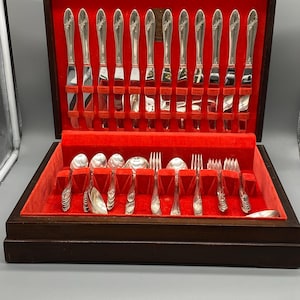 May include: A wooden flatware chest with a bright red velvet interior containing a set of silver-toned knives, spoons, and forks. The knives are arranged vertically in the lid, while the spoons and forks are organized in the base slots.