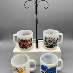 May include: Four white ceramic mugs with various floral and fruit designs, including strawberries, daisies, and blue flowers. The mugs are displayed on a white wooden stand with a black metal rack.