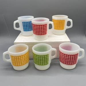 Puede incluir: Seis tazas de cerámica blanca con asa. Cada taza presenta un estampado de cuadros de diferentes colores: azul, rojo, amarillo, naranja, verde y rojo. Las tazas están dispuestas sobre una superficie blanca sobre un fondo gris.