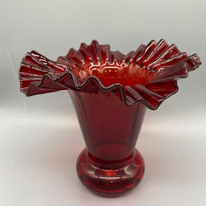 Peut inclure: Un vase en verre rouge profond doté d'une large ouverture évasée avec un bord décoratif à volants et froncé. Le corps du vase est lisse et se rétrécit vers une base circulaire arrondie, mettant en valeur une couleur cramoisie translucide.
