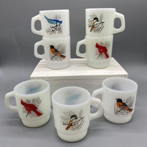 Puede incluir: Un conjunto de tazas de cerámica blanca con ilustraciones de aves. Cada taza, con asa, presenta un diseño diferente, como un arrendajo azul, un cardenal y un bolsero. Las tazas están apiladas y dispuestas sobre una superficie blanca.