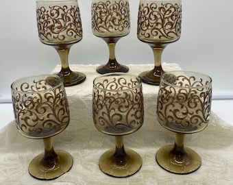Könnte beinhalten: Sechs Vintage-Weingläser mit braunen Stielen und Böden. Die klaren Glasschalen sind mit einem braunen, wirbelnden, verzierten Muster verziert. Die Gläser sind auf einer weißen Spitzentischdecke angeordnet und schaffen eine klassische und elegante Ästhetik.