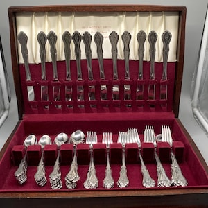 May include: A wooden storage chest containing a silver-plated flatware set. The interior features a cream-coloured satin lining with the text 1847 ROGERS BROS and a deep red velvet base with individual slots for knives, spoons, and forks.