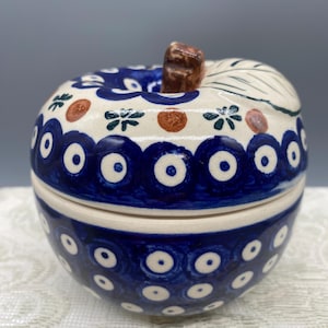 Può includere: Un barattolo in ceramica a forma di mela con coperchio su base color crema, decorato con un motivo dipinto a mano di cerchi blu scuro con centri bianchi. Il coperchio presenta un picciolo marrone, una foglia verde, punti arancioni e motivi floreali.