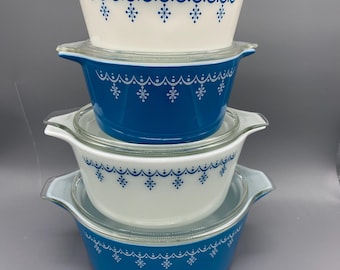 Op de afbeelding: Een stapel van vier ronde glazen ovenschaaltjes met doorzichtige glazen deksels. De schaaltjes wisselen af tussen wit en blauw, elk voorzien van een blauw of wit decoratief patroon van geschulpte lijnen en sterretjes rond de bovenrand.