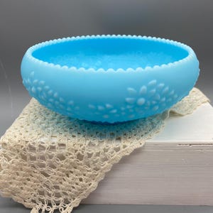May include: A bright blue milk glass bowl featuring a scalloped rim and a raised floral pattern around the exterior. The bowl has a smooth, opaque finish and a rounded shape, showcasing a consistent light blue colour throughout the glass surface.
