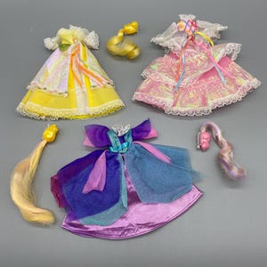 May include: Three doll dresses in yellow, pink, and purple with matching hair pieces. The dresses have layered skirts, lace trim, and colorful ribbon accents. The hair pieces are blonde and pink.