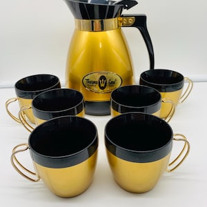 Op de afbeelding: Een vintage gouden en zwarte Thermo Serv geïsoleerde serveerset. De set bevat een gouden karaf met een zwart deksel en handvat, en zes bijpassende kopjes. De kopjes hebben gouden bodems en zwarte interieurs. De karaf heeft het opschrift "Thermo Serv".