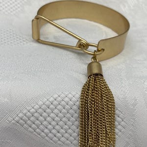 May include: A gold-colored metal bracelet with a safety pin-style clasp and a dangling tassel. The tassel is made of small gold chains. The bracelet is on a white, textured surface.