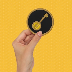 May include: A black and gold embroidered patch featuring a banjo with a yellow background.
