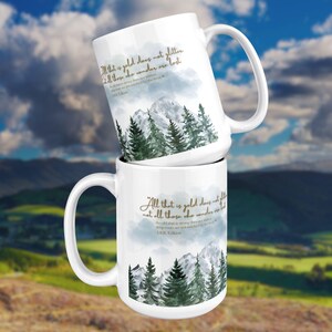 May include: Two white ceramic mugs stacked, each featuring a mountain and forest scene with a quote in gold lettering. The quote reads "All that is gold does not glitter, not all those who wander are lost." by J.R.R. Tolkien.