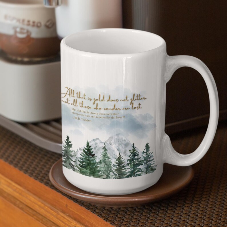 May include: White ceramic mug with a quote from J.R.R. Tolkien, featuring a mountain and forest scene. The quote reads, "All that is gold does not glitter, not all those who wander are lost." The mug is on a brown coaster.