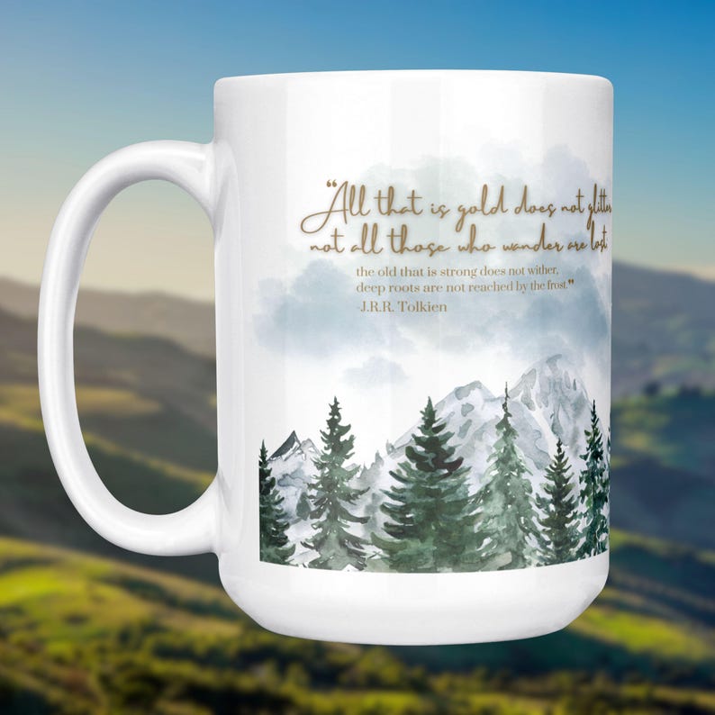 May include: White ceramic mug featuring a mountain and forest scene with the quote "All that is gold does not glitter, not all those who wander are lost" by J.R.R. Tolkien. The mug has a large handle.