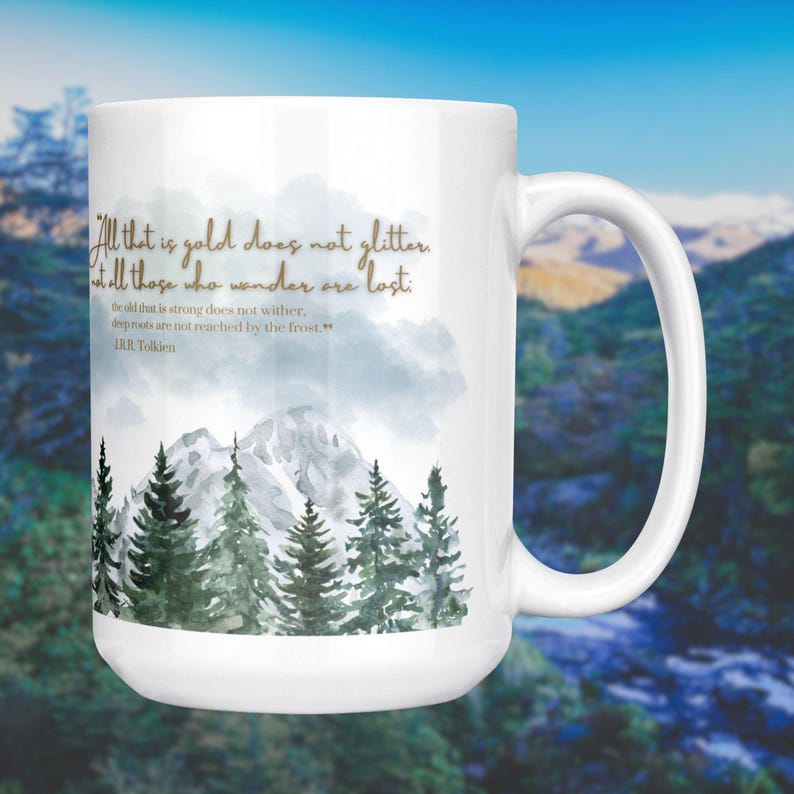 May include: White ceramic mug featuring a quote from J.R.R. Tolkien: "All that is gold does not glitter, not all those who wander are lost." The design includes a watercolor illustration of a mountain range and evergreen trees.