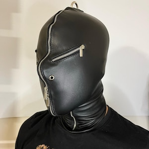 May include: Black leather hood with a full face zipper and a metal ring at the top. The hood covers the head and neck, with a zipper closure around the face. The design is sleek and minimalist.