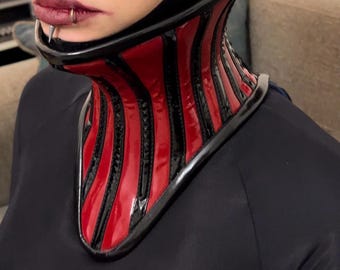 May include: A person wearing a black latex hood and a red and black corset with a pointed collar.