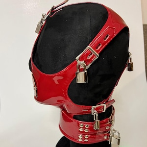 以下が含まれることがあります： シルバーの南京錠とバックルが付いた、赤色の光沢のある革製フード。 フードは頭と首を覆い、ネックバンドとストラップが付いています。 製品は黒いマネキンの頭に表示されています。