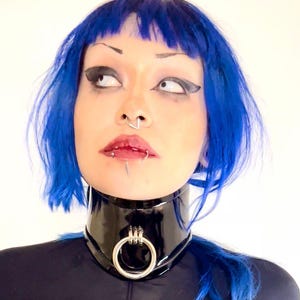 May include: A black, high-collared choker with a large silver ring detail. The choker is made of a shiny material and is worn with a black long-sleeved top. The person in the image has blue hair and facial piercings.