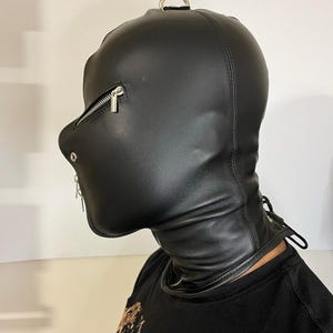 May include: Black leather hood with a zippered mouth and eye openings. The hood features a metal D-ring at the top and a zipper closure on the side. The neck area has a lace-up design for adjustment.