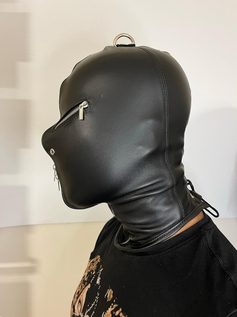May include: Black leather hood with a zippered mouth and eye openings. The hood features a metal D-ring at the top and a zipper closure on the side. The neck area has a lace-up design for adjustment.