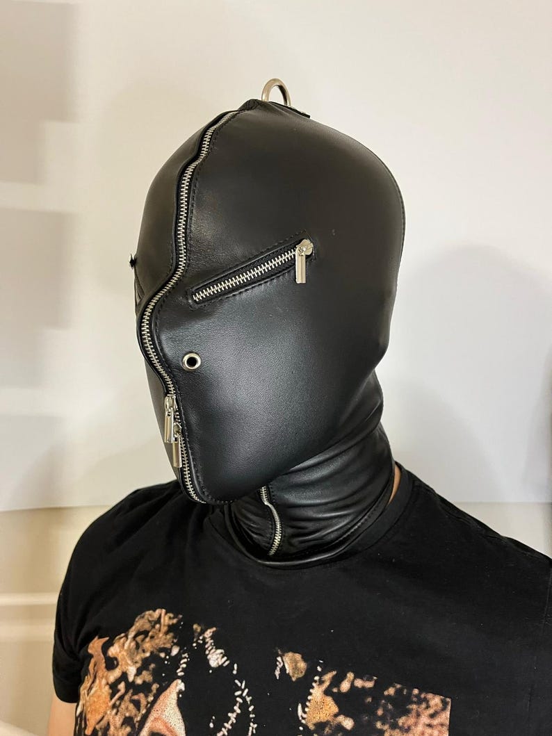 May include: Black leather hood with a full face zipper and a metal ring at the top. The hood covers the head and neck, with a zipper closure around the face. The design is sleek and minimalist.