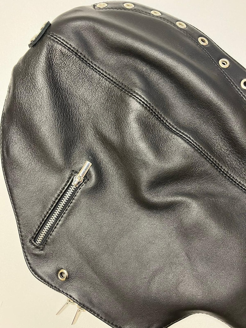 May include: Close-up of a black leather accessory with a zipper and metal grommets. The accessory has a textured surface and is stitched with a visible seam. Silver-colored metal spikes are visible at the bottom.