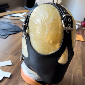 May include: Black leather mask with a cutout for the nose and mouth. The mask is attached to a black leather harness with silver buckles and rings.